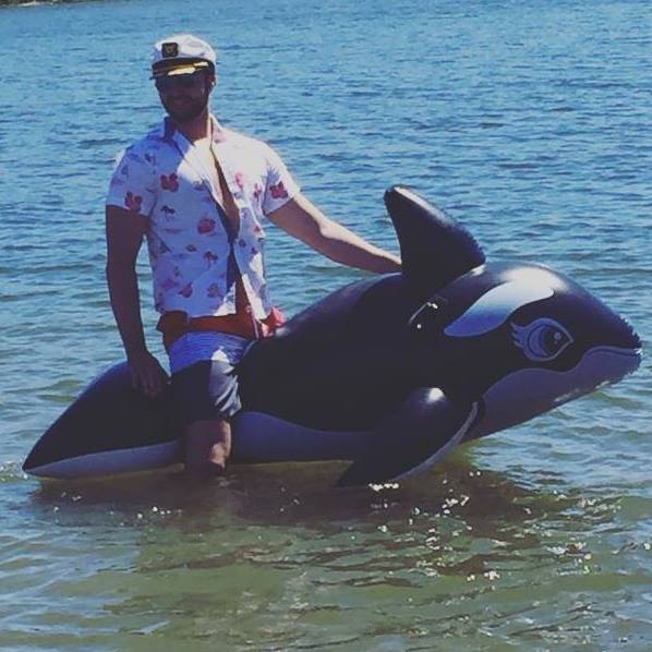 Travis Jones standing in water with inflatable orca wearing a captain hat on a beach day